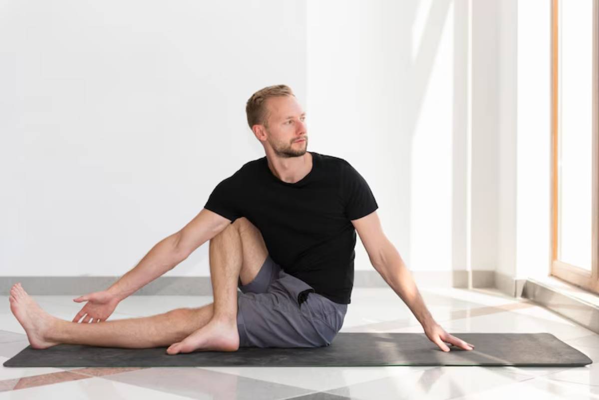 A person sits on a yoga mat in a bright room, performing a seated stretch with one arm extended and one leg relaxed.