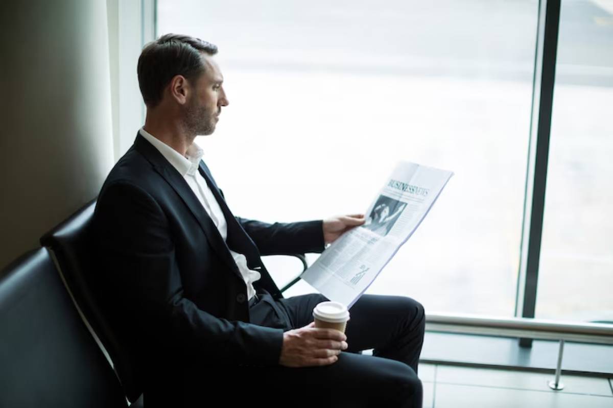 A man in a black suit sits in a modern setting, reading a newspaper and holding a coffee cup, with large windows in the background.