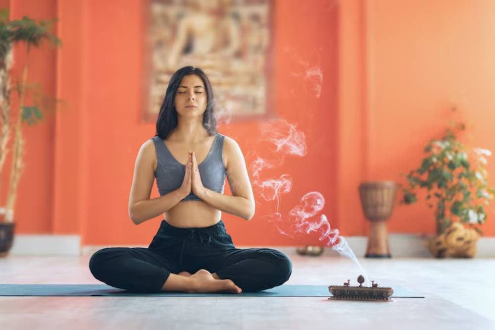 A woman sits in a meditative pose on a yoga mat, surrounded by a warm orange backdrop and fragrant incense smoke curling in the air.