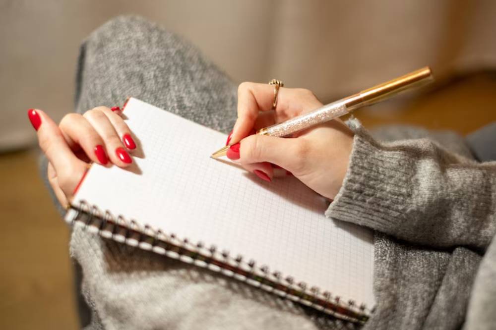 A close-up of a person's hands with red nails writing in a grid notebook using a glittery gold pen while wearing a grey sweater.