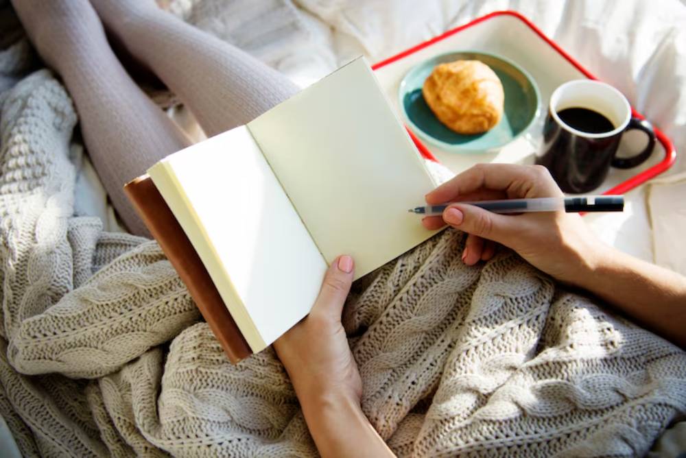 A person writes in a blank notebook while cozying up in a knitted blanket, with a croissant and coffee on a tray nearby.