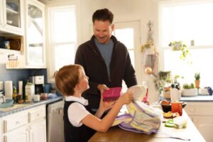 A child holds a lunchbox while an adult prepares a backpack in a bright, cozy kitchen filled with plants and kitchenware.