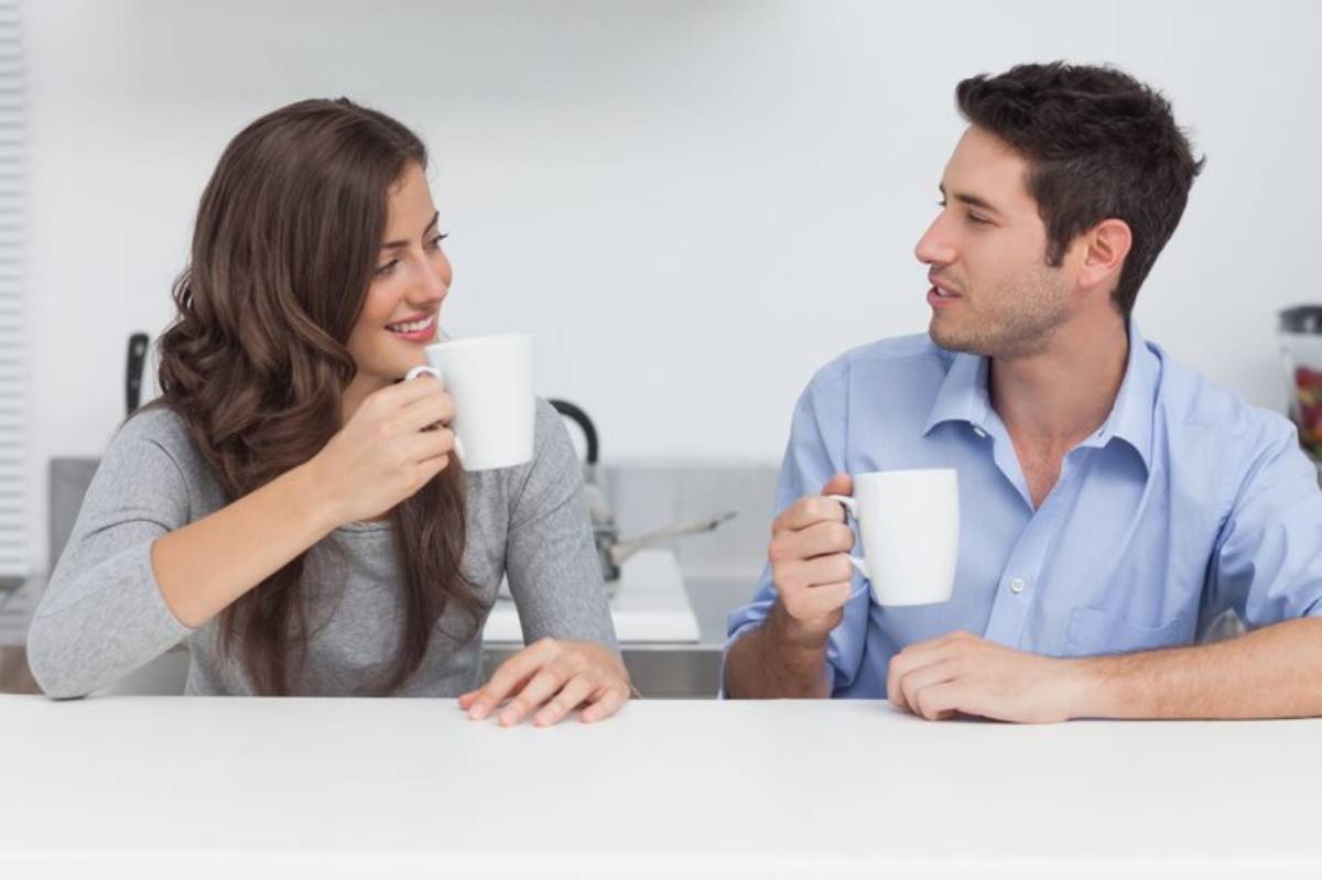 Two people sit at a white table, each holding a white mug, engaged in conversation in a bright kitchen setting.