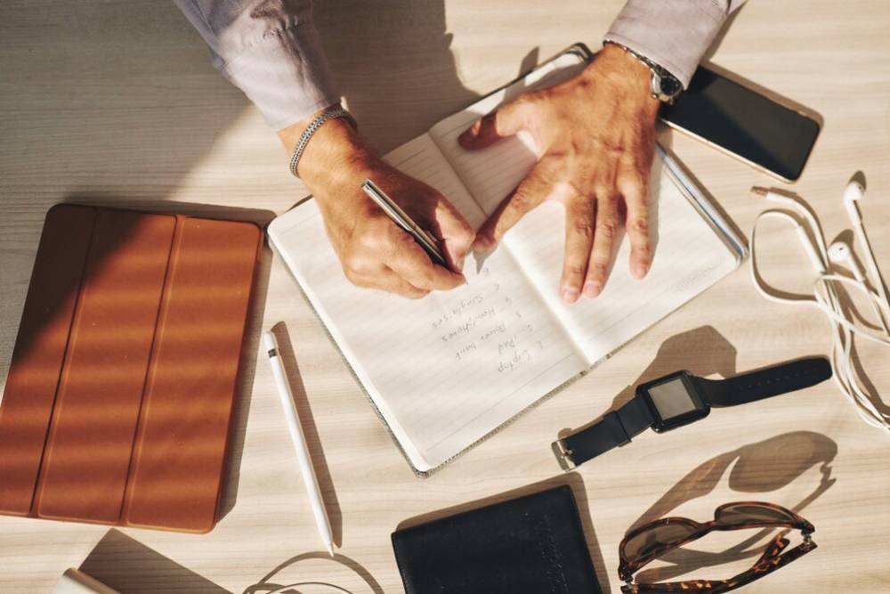 A person's hand holds a pen, writing in a notebook surrounded by a tablet, phone, sunglasses, and a smartwatch on a wooden desk.