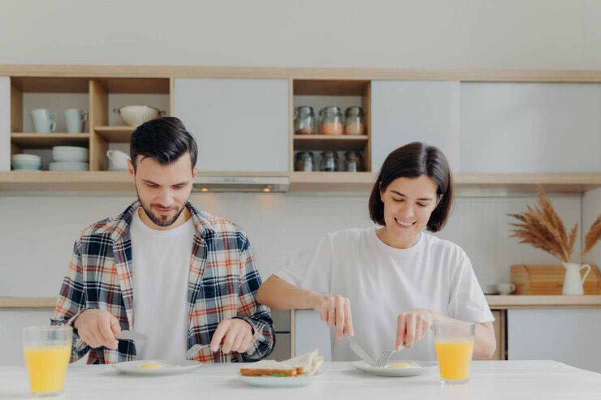 Two people sitting at a table, focused on their plates, with a glass of orange juice nearby and a minimalistic kitchen in the background.