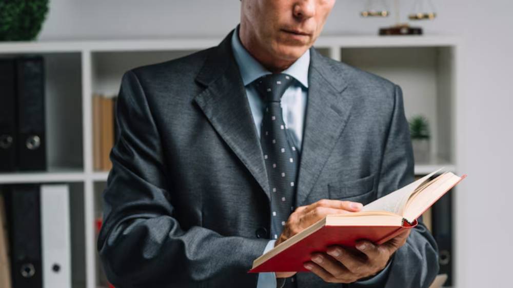 A man in a suit reads a book attentively, standing in an office with shelves of files and decorative plants in the background.