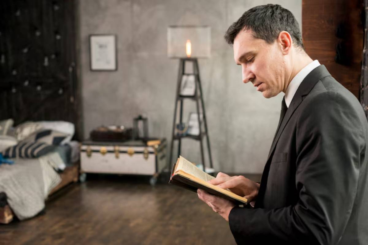 A man in a suit is reading a book in a stylish room featuring a bed with patterned linens and a contemporary lamp.