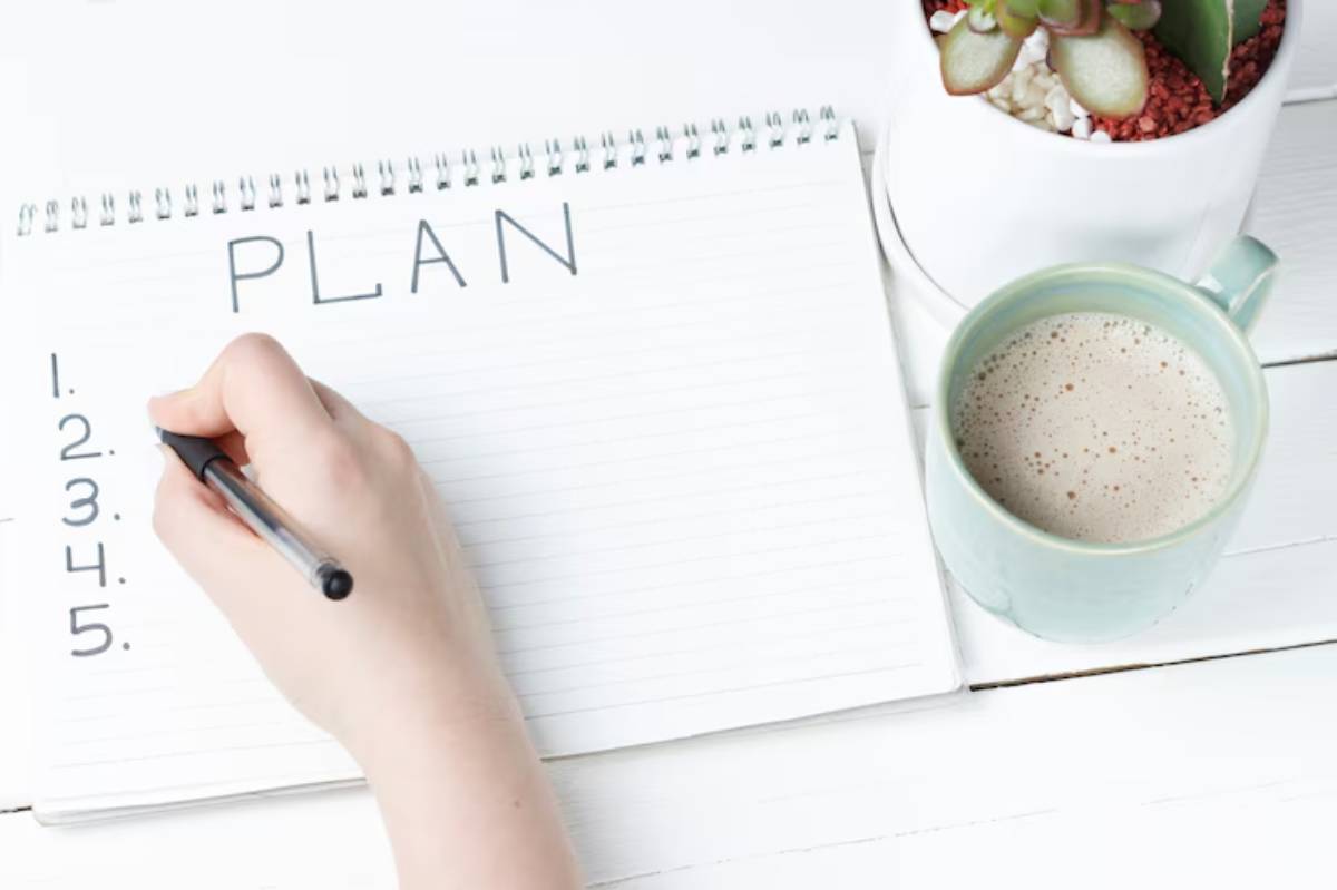 A hand writes a list titled 'PLAN' in a notebook beside a cup of coffee and a potted plant on a white wooden table.