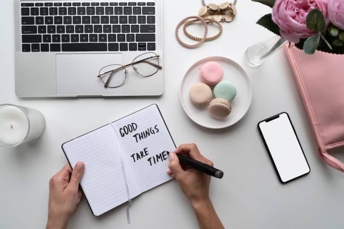 A hand writes 'GOOD THINGS TAKE TIME' in a notebook, surrounded by a laptop, macarons, glasses, and a pink bag on a white desk.