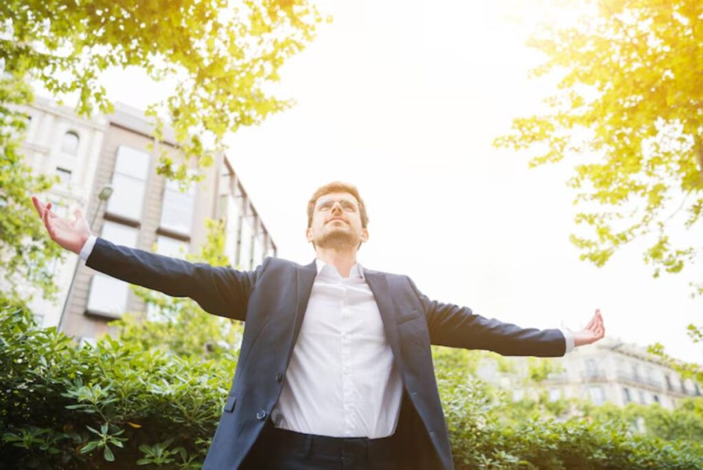 A man in a suit stands with arms outstretched, basking in sunlight amidst greenery and city buildings, conveying gratitude.