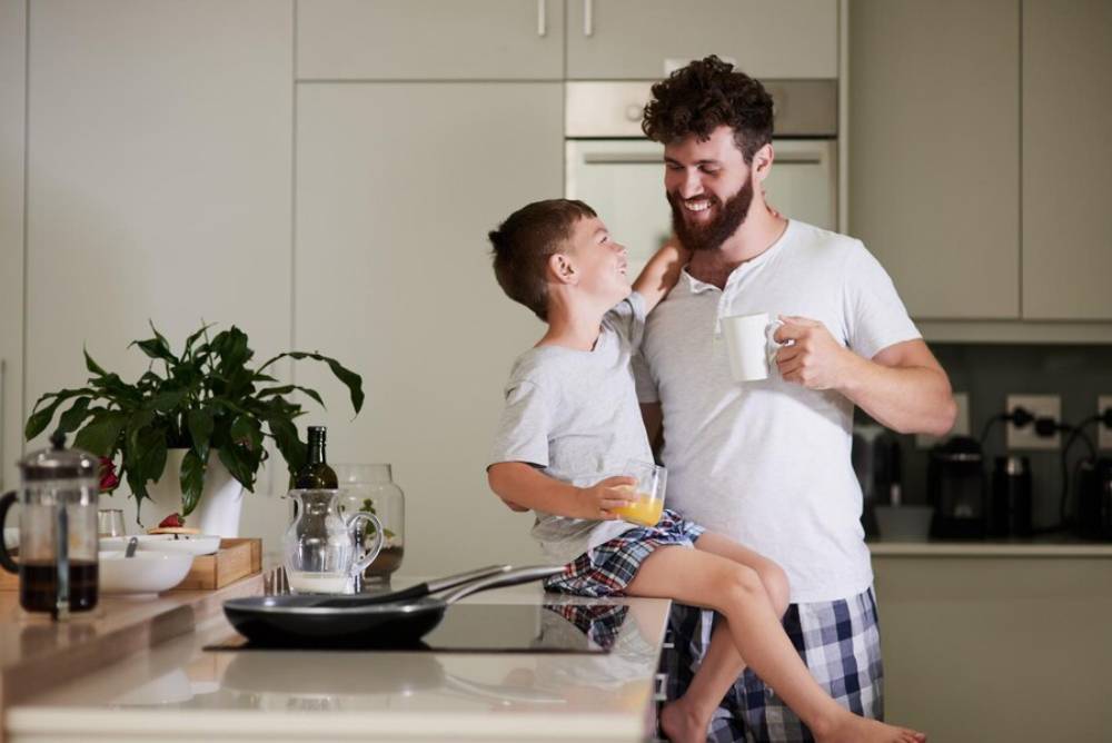 A father holds a cup while his son sits on the kitchen counter, enjoying a glass of juice amidst a cozy breakfast setting.