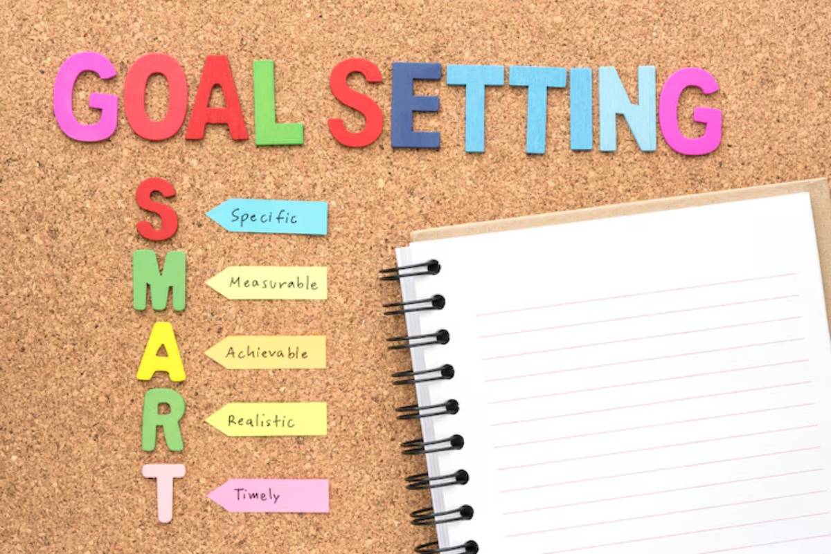 Colorful letters spell 'Goal Setting' on a corkboard, with the SMART criteria listed and a blank notepad beside them.