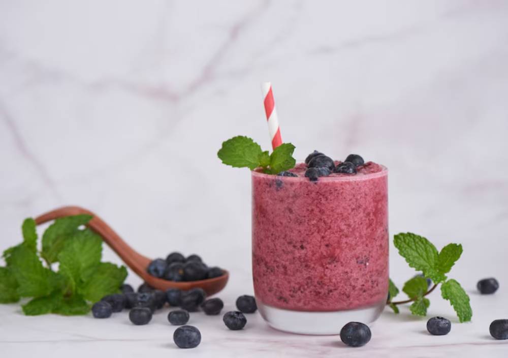 A refreshing blueberry smoothie in a glass with a red straw, garnished with mint leaves, surrounded by blueberries and mint. 
