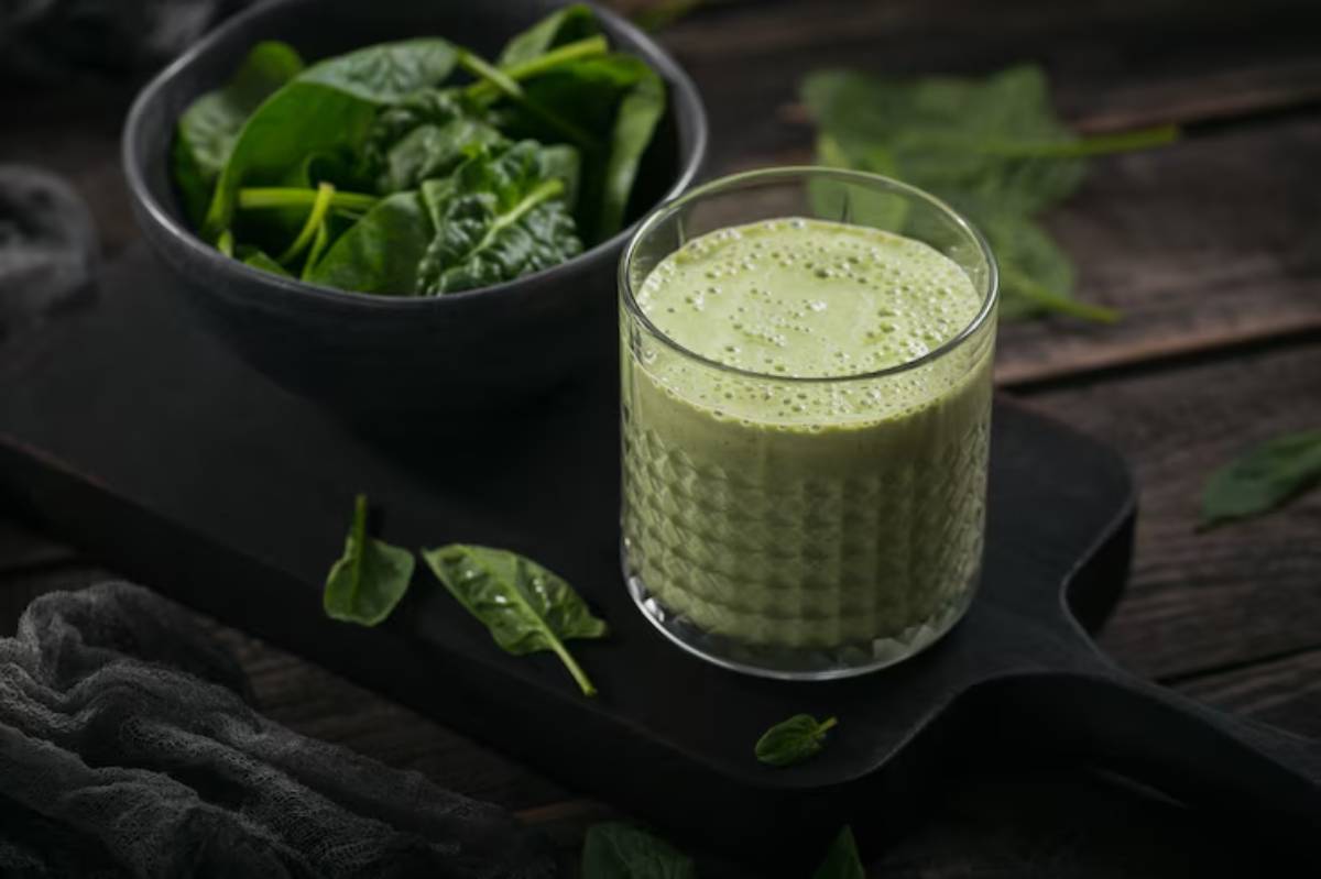 A glass of green smoothie beside a bowl of fresh spinach leaves, set on a dark wooden surface with scattered spinach.
