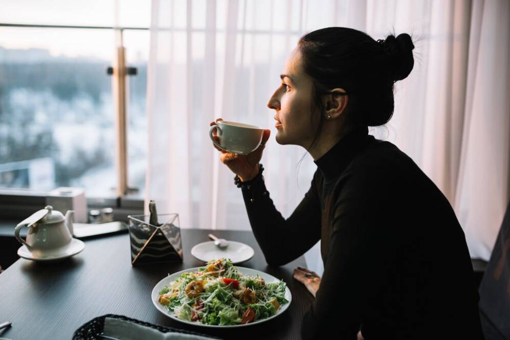 A woman in a black turtleneck enjoys a cup of tea beside a fresh salad, with a city view through large windows.