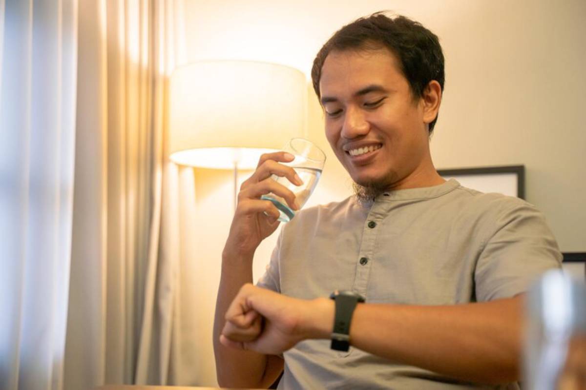 A person sits at a table, holding a glass of water, while checking a smartwatch on their wrist, with soft lighting in the background.