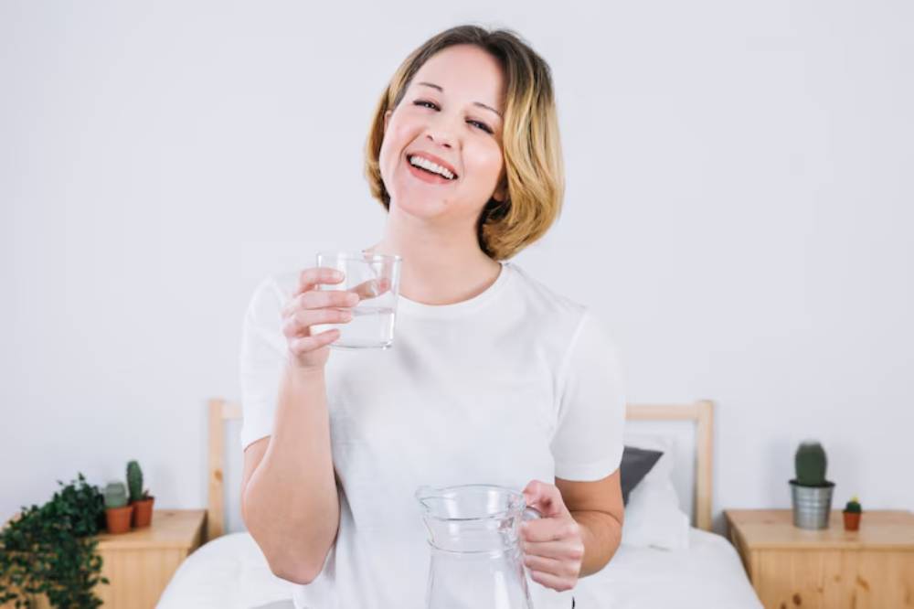 A person in a white shirt holds a glass of water in one hand and a pitcher of water in the other, standing in a bright, minimalistic bedroom.
