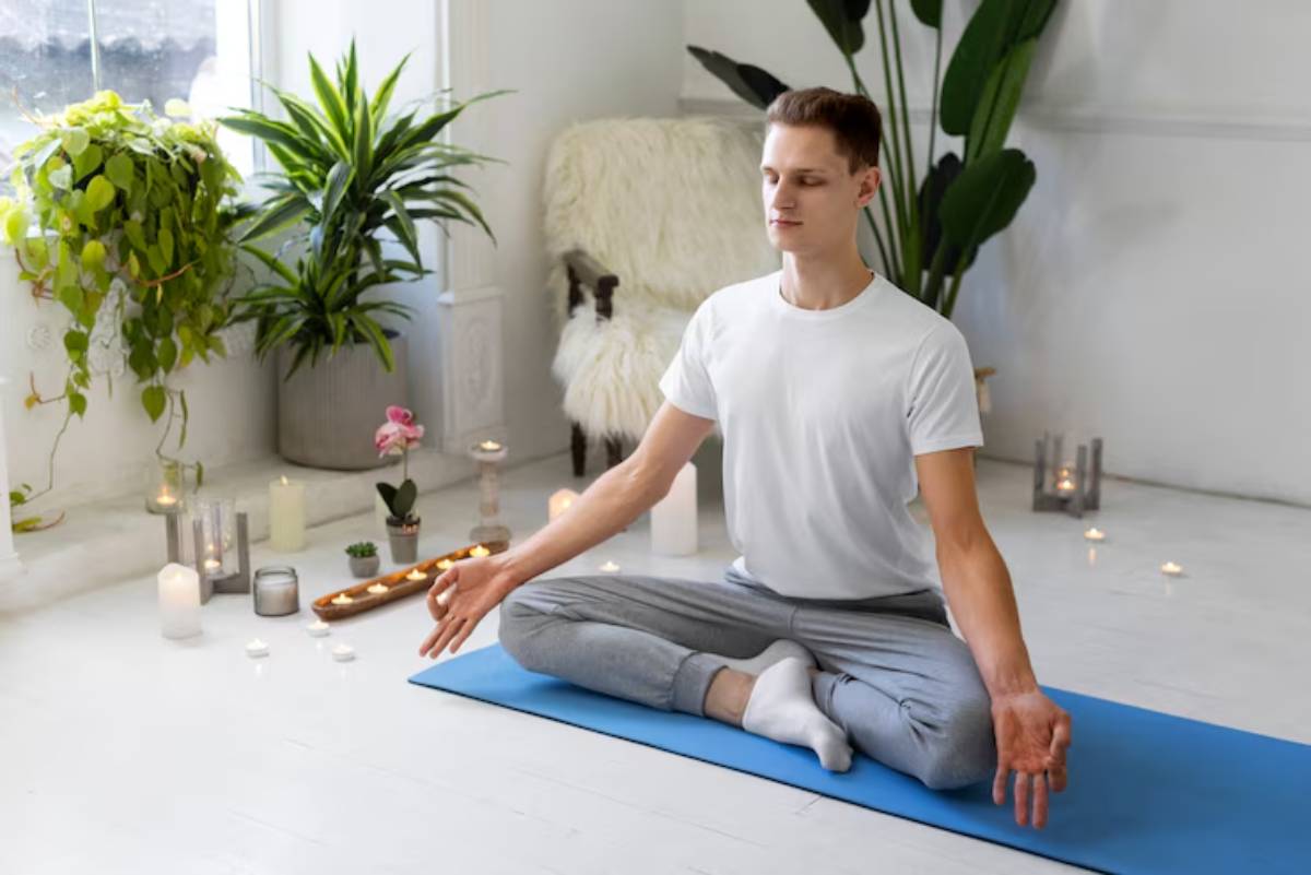 A person meditating on a blue yoga mat, surrounded by plants and candles in a bright, serene indoor space.