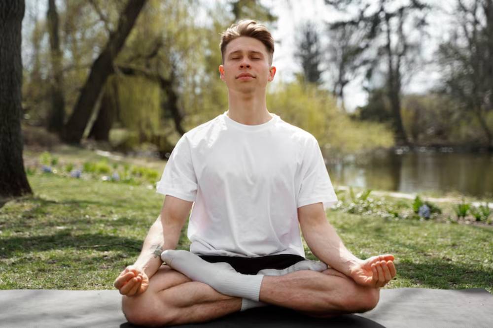 A person sits cross-legged on a yoga mat by a serene lake, practicing meditation amidst lush greenery and soft sunlight.
