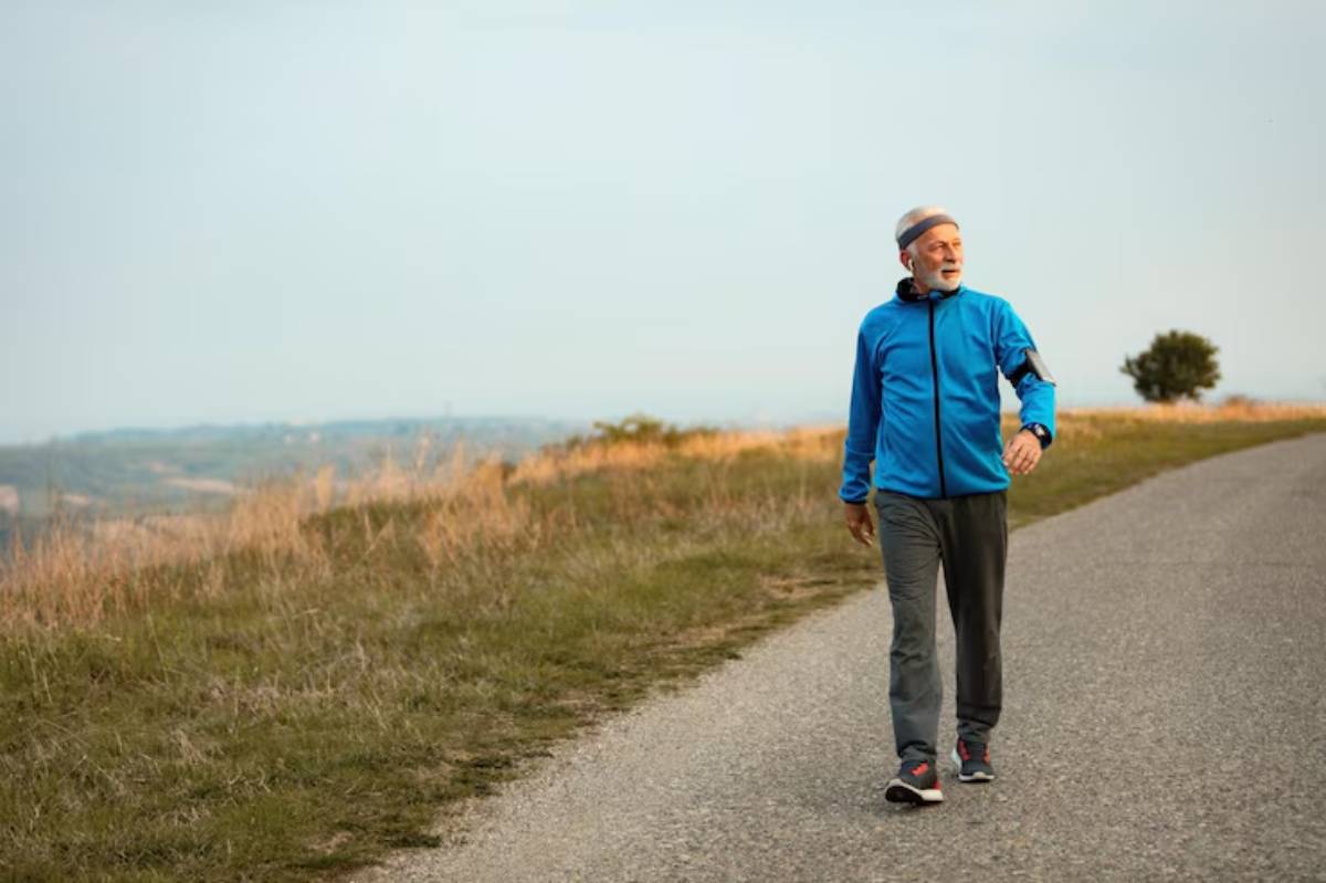 A person in a blue jacket and gray pants walks along a scenic path with rolling hills in the background under a clear sky.