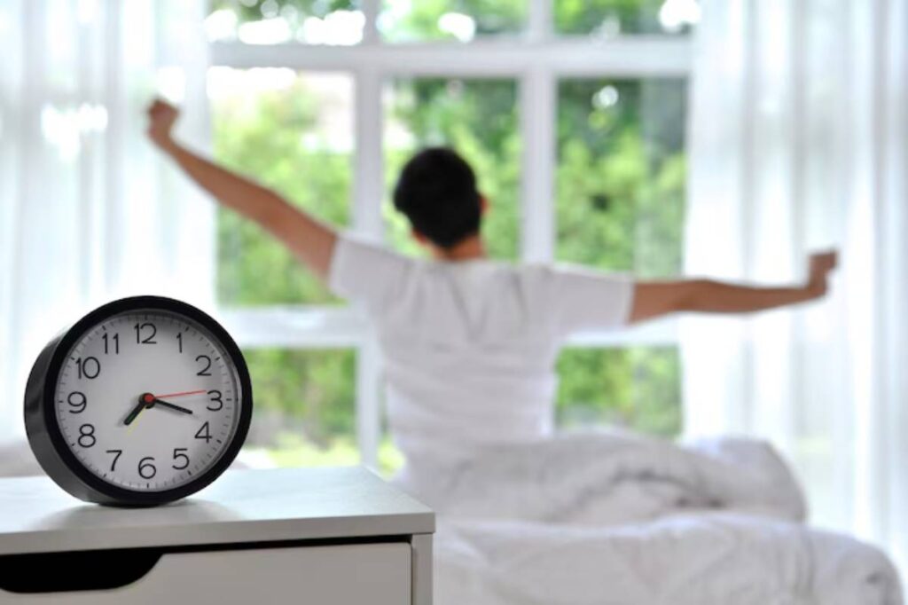 A man stretches in bed by a window, while a clock on a nightstand shows the time. Bright light filters through sheer curtains.