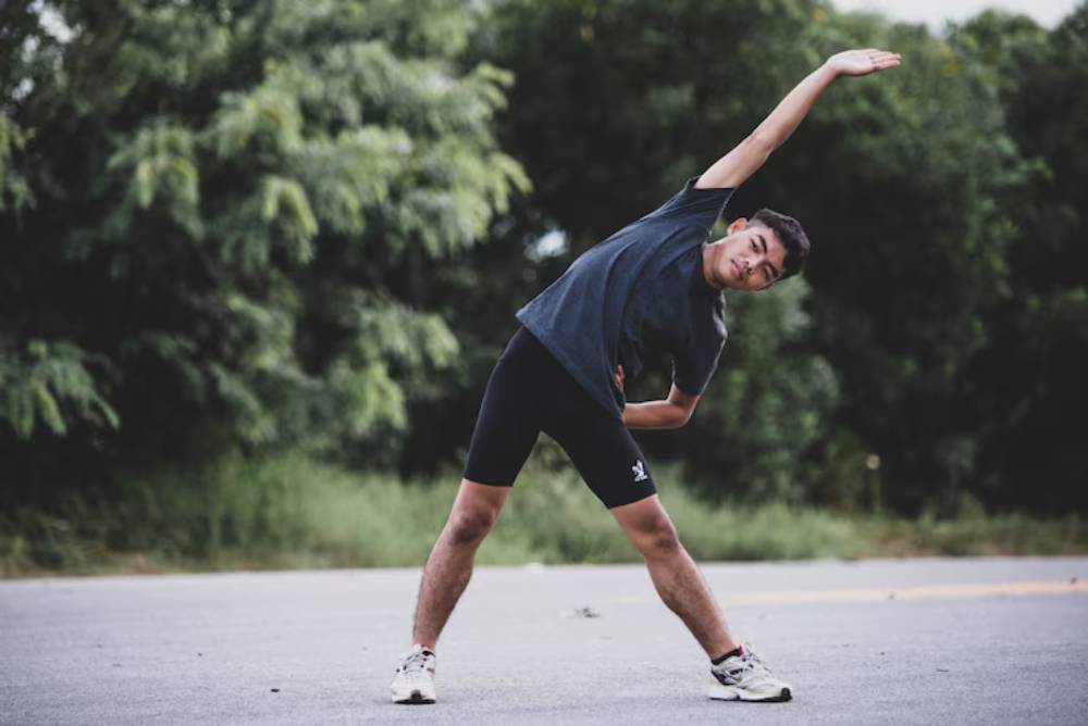 A person in a gray t-shirt and black shorts stretches sideways on a road, surrounded by lush greenery.