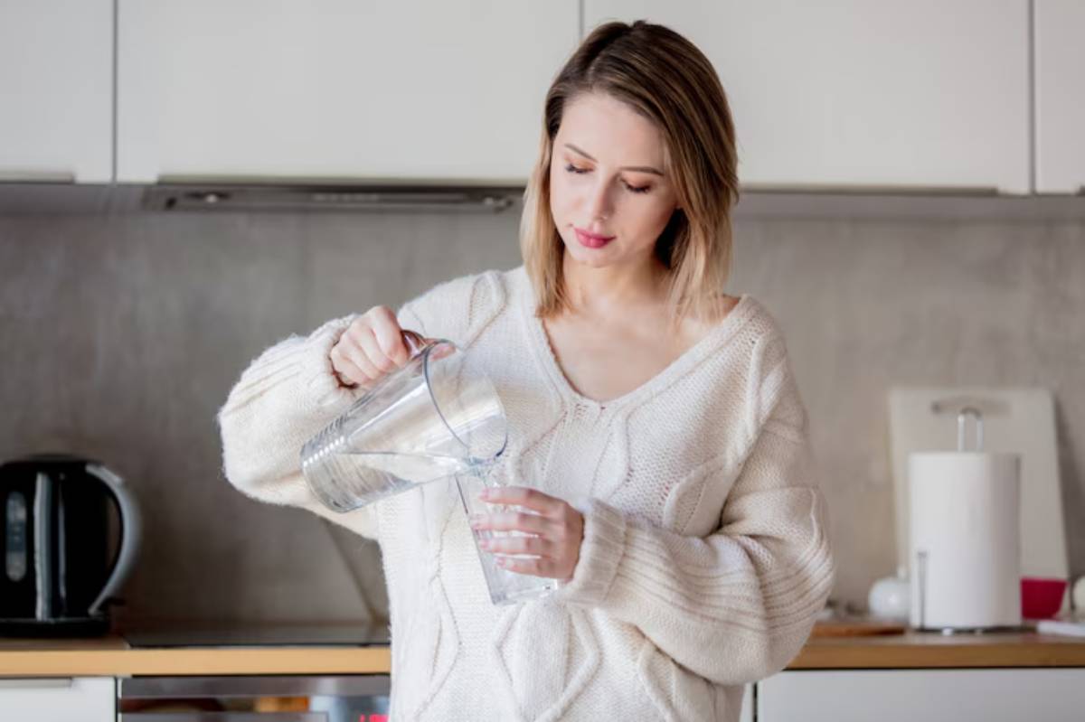 A person in a cozy sweater pours water from a pitcher into a glass in a modern kitchen, with a kettle and kitchen supplies in the background.