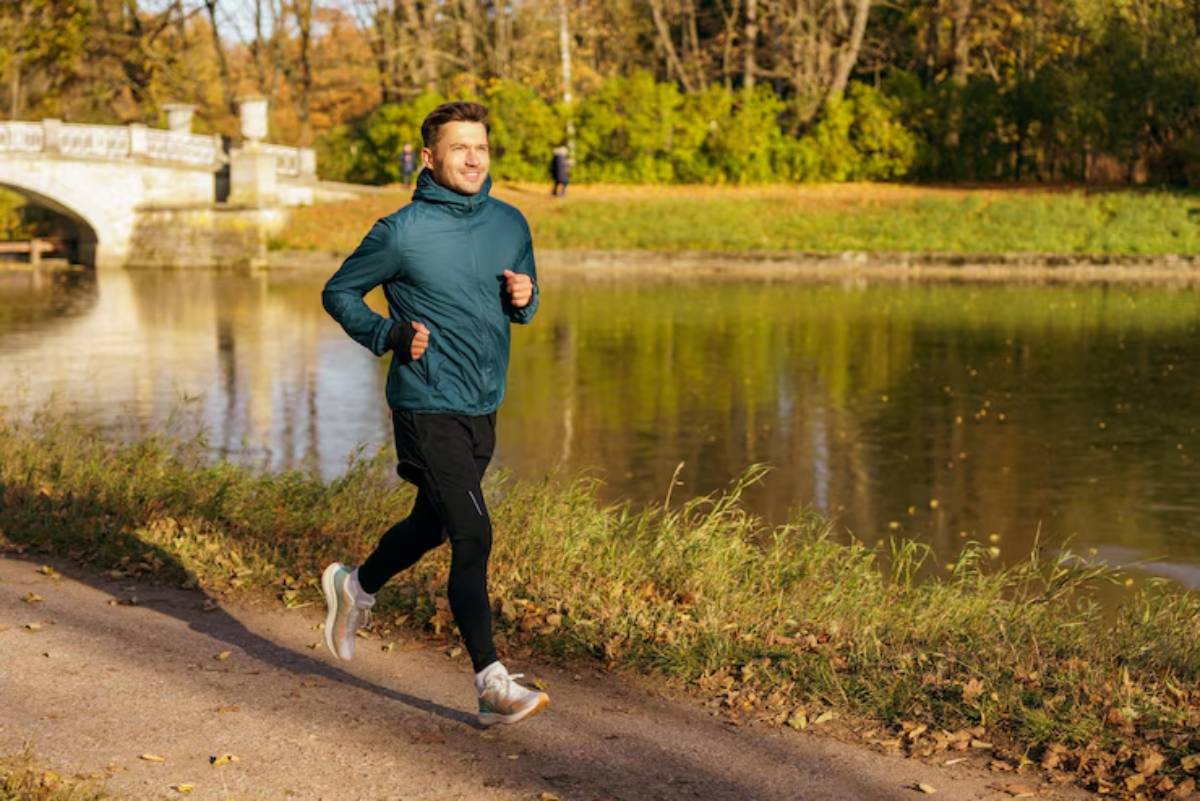 A jogger in a blue jacket runs along a path next to a calm river, surrounded by autumn foliage and trees. 