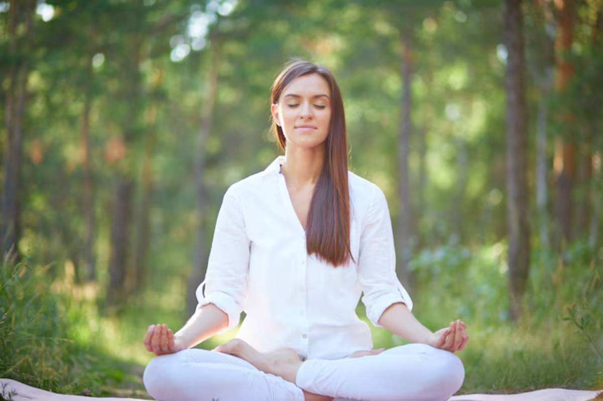A woman in a white shirt meditates peacefully on a mat in a serene forest, surrounded by green trees and soft sunlight.