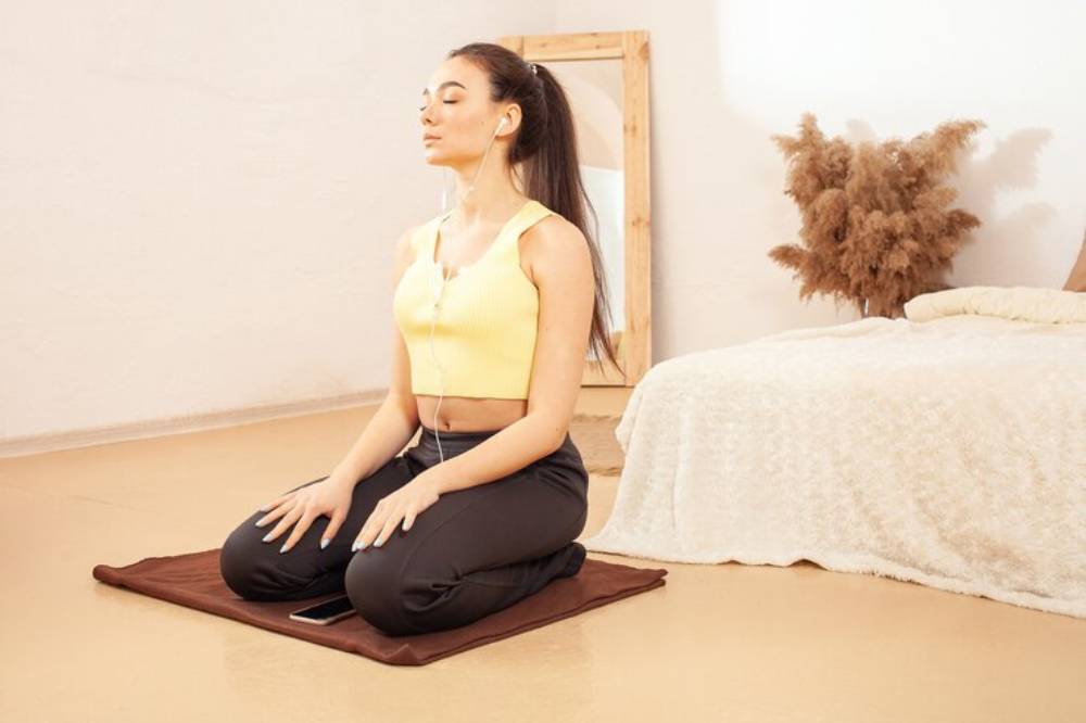 A person sits cross-legged on a mat, wearing comfortable workout clothes, with a serene atmosphere created by a soft rug and plants.