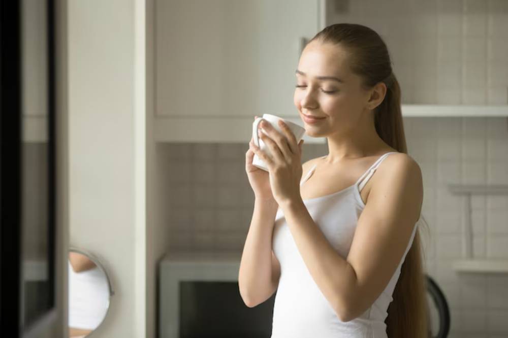 A woman in a white tank top holds a cup while standing in a bright kitchen, surrounded by minimalist decor and appliances.