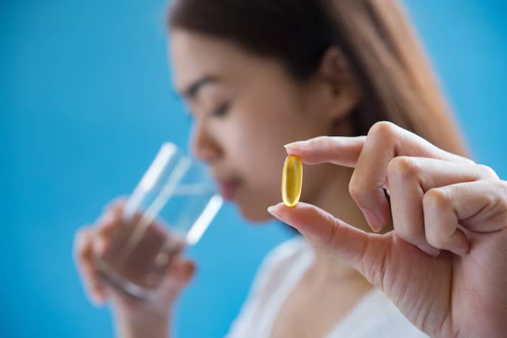A person holds a glass of water in one hand while displaying a supplement capsule in the other, set against a blue background.
