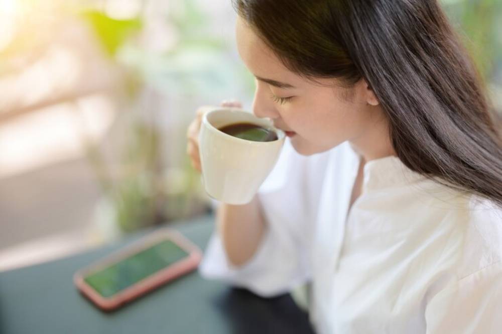 A person holds a white cup of coffee, sitting at a table with a smartphone nearby, surrounded by greenery and soft sunlight.