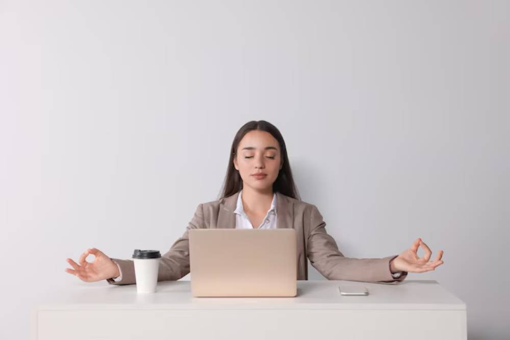 A person in a suit sits at a desk with a laptop, coffee cup, and smartphone, practicing meditation with hands in a mudra pose.