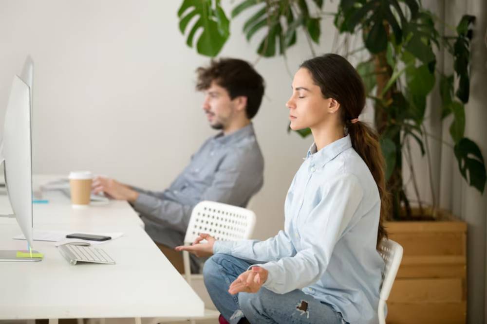 A woman practices mindfulness meditation at a modern workspace, while a man sits at a desk working on a computer. Green plants are visible.