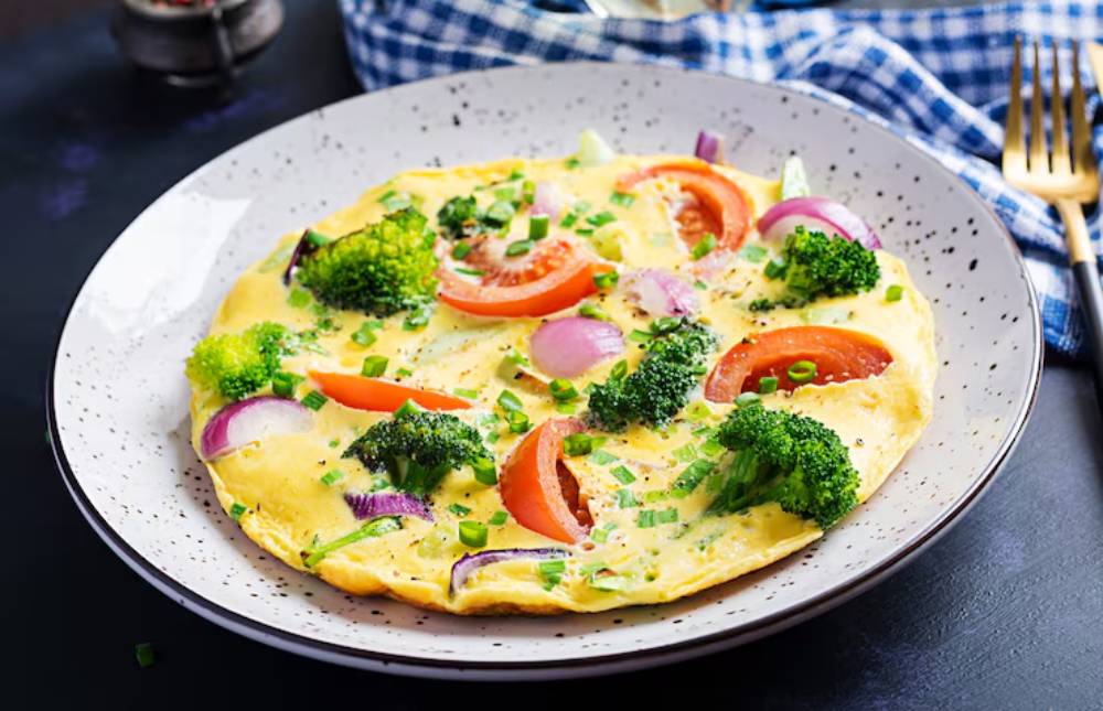 A delicious vegetable omelette topped with broccoli, tomatoes, and red onions, garnished with green onions on a speckled plate. 