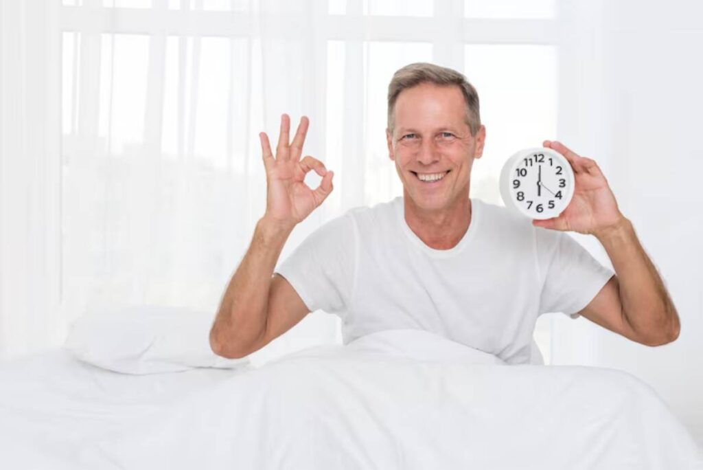A man in a white t-shirt sits in bed, holding a clock in one hand and making an 'OK' gesture with the other. Light fills the room.