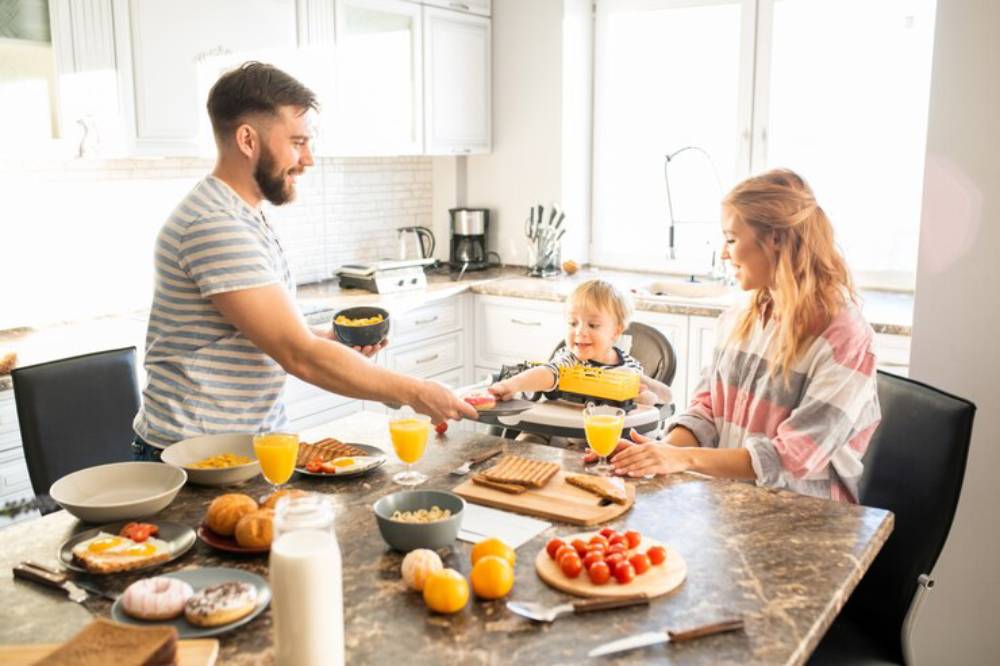 A family enjoys breakfast at a modern kitchen table, with various dishes including eggs, pastries, and orange juice.