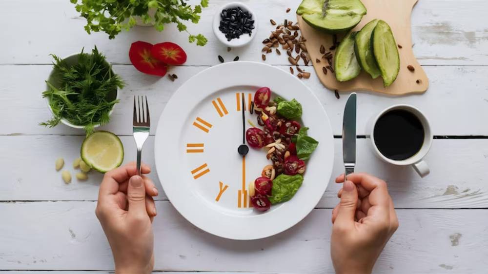 A plate shaped like a clock features a colorful salad, with hands holding a fork and knife, surrounded by fresh ingredients and coffee.
