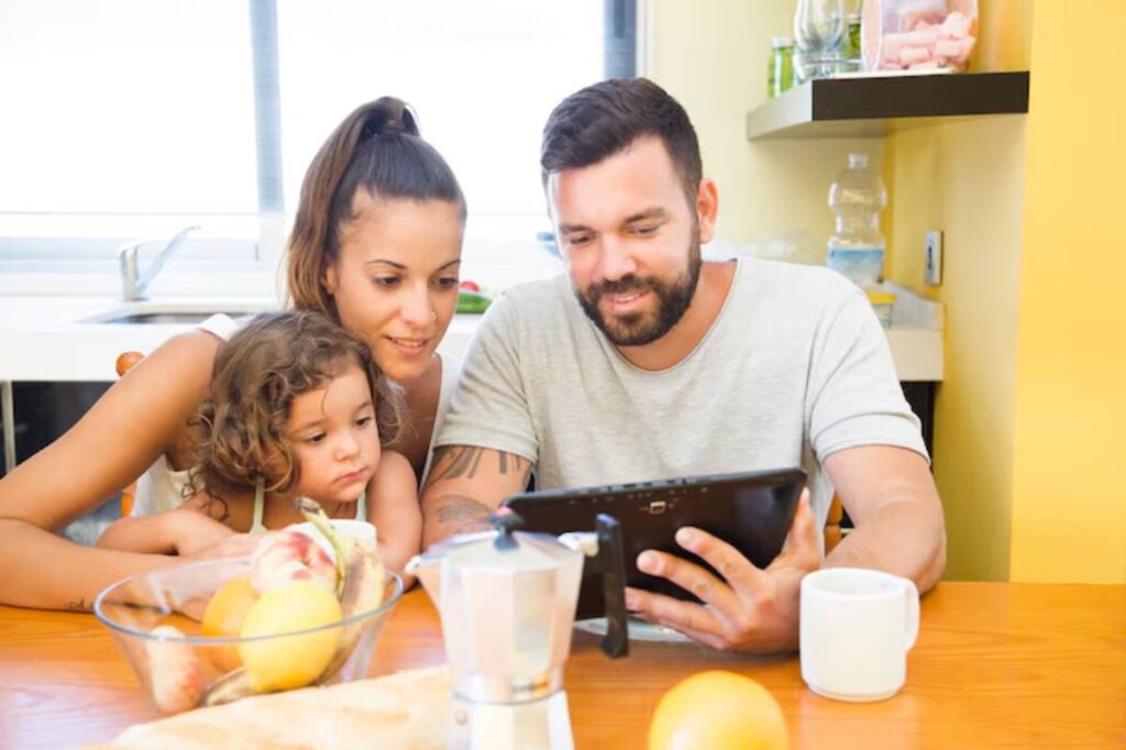A family sits at a wooden dining table, engaged with a tablet. Fresh fruits and a coffee maker are visible in the foreground.