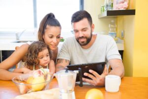 A family sits at a wooden dining table, engaged with a tablet. Fresh fruits and a coffee maker are visible in the foreground.