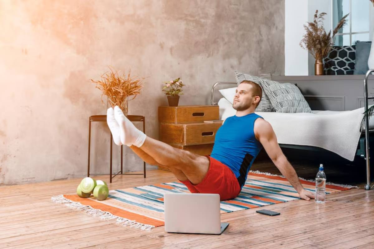 A person exercises at home on a colorful rug, with dumbbells, water bottle, and a laptop in a cozy living space.