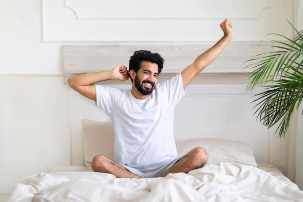 A person in a white t-shirt stretches while sitting cross-legged on a bed, surrounded by soft bedding and a potted plant.