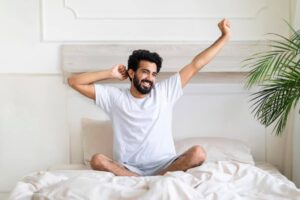 A person in a white t-shirt stretches while sitting cross-legged on a bed, surrounded by soft bedding and a potted plant.