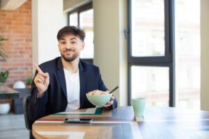 A person in a dark blazer sits at a wooden table, holding a bowl of cereal, gesturing with one hand, with a drink beside them.