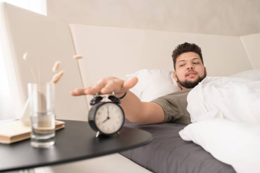 A person reaching for an alarm clock on a bedside table while lying in bed, surrounded by white bedding and soft lighting.