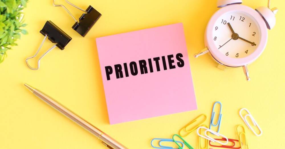 A pink sticky note labeled 'PRIORITIES' surrounded by paper clips, a gold pen, black binder clips, and a vintage alarm clock on a yellow background.