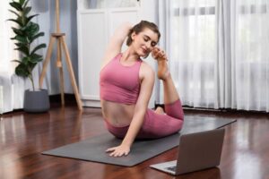A person in sporty attire practices yoga on a mat, with a laptop nearby, in a sunlit room filled with greenery.