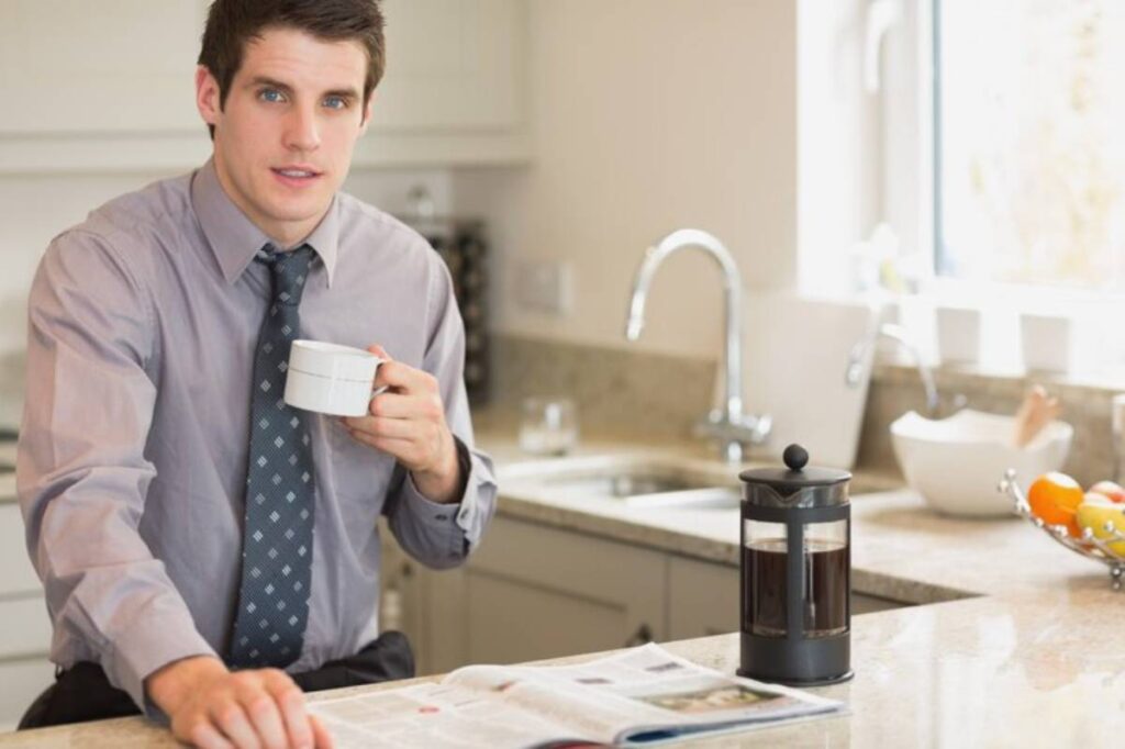 A man in a dress shirt and tie sits at a kitchen counter, sipping coffee, with a French press and a magazine in front of him.