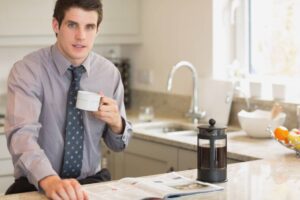 A man in a dress shirt and tie sits at a kitchen counter, sipping coffee, with a French press and a magazine in front of him.